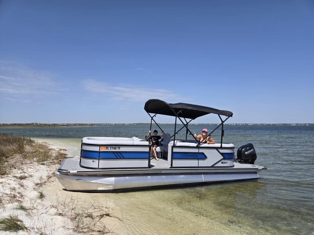 Woman and child on a Pensacola Beach pontoon rental anchored on white sand under a clear blue sky.