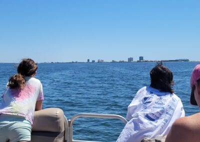 Two passengers on the back of a luxury tritoon spotting a wild dolphin fin in the water, with the Pensacola Beach skyline in the background during a captained boat tour.
